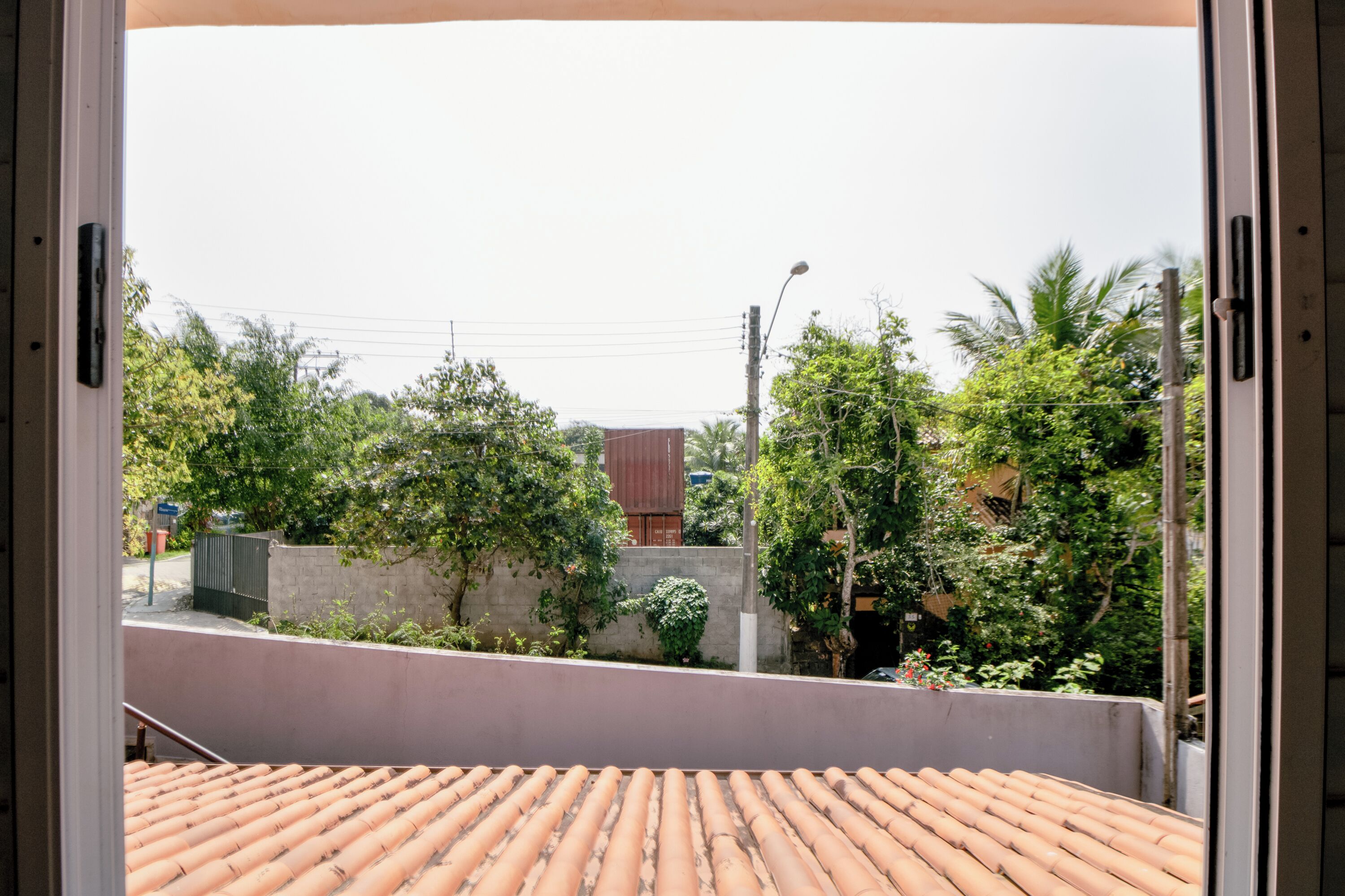 Vista desde un balcón con suelo de baldosas, pared de hormigón y vegetación, incluyendo árboles y una valla.