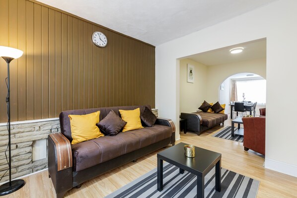 A modern living room with a brown leather sofa, a coffee table, and a lamp. There is a clock on the wall and a dining area in the background. - 2-bed House - Sleeps 7 & Free Parking (Barking)