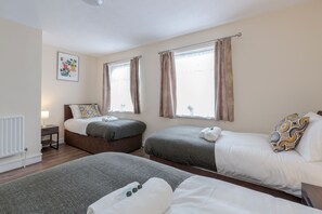 A hotel room with two single beds, a small bedside table with a lamp, a radiator, and a window with curtains. - 2-bed House - Sleeps 7 & Free Parking (Barking)