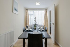 A dining area with a table set for four, chairs, a potted plant, and a window with curtains. - 2-bed House - Sleeps 7 & Free Parking (Barking)