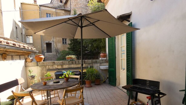 Une terrasse extérieure avec une table ronde, des chaises, un grand parasol, des plantes en pot et un barbecue.