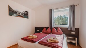 A double bed with red pillows and towels, a window with curtains, a small bedside table, and a painting on the wall. - Fankhauser by Interhome (Kaprun)