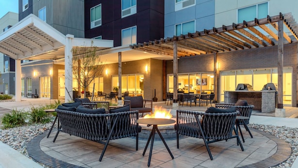 An outdoor patio area with a round table and wicker chairs, a fire pit, and a pergola. - TownePlace Suites by Marriott Chesterfield (New Baltimore)