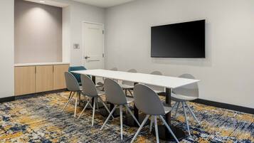 A conference room with a white table, grey chairs, a flat-screen TV on the wall, and a patterned carpet.