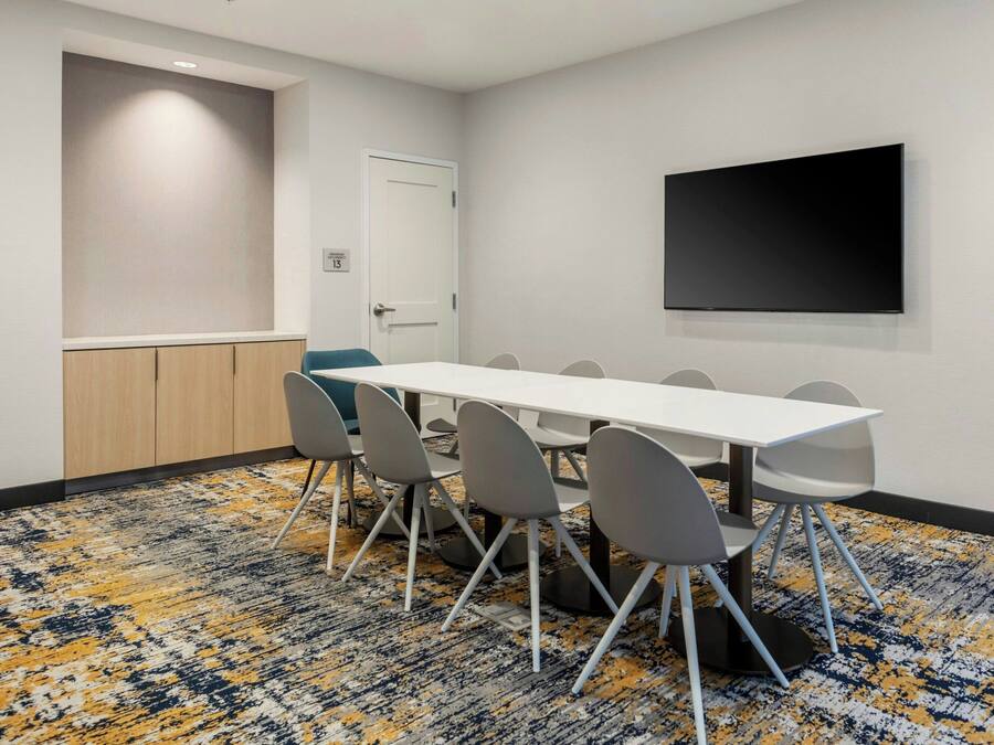 A conference room with a white table, grey chairs, a flat-screen TV on the wall, and a patterned carpet.