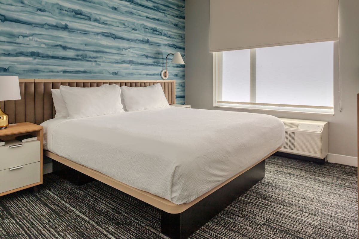A hotel room with a bed, a nightstand, a lamp, a window, and a wall with a blue pattern.