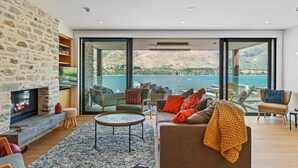 A modern living room with a stone wall, a fireplace, and a large window overlooking a lake. - Luxury Lake View (Wānaka)