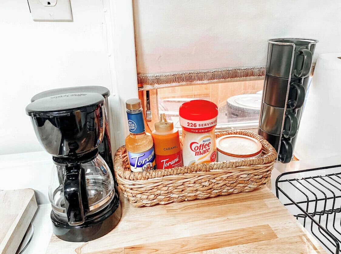 A kitchen counter with a coffee maker, a basket containing Torani syrup, a jar of coffee creamer, and a coffee machine.