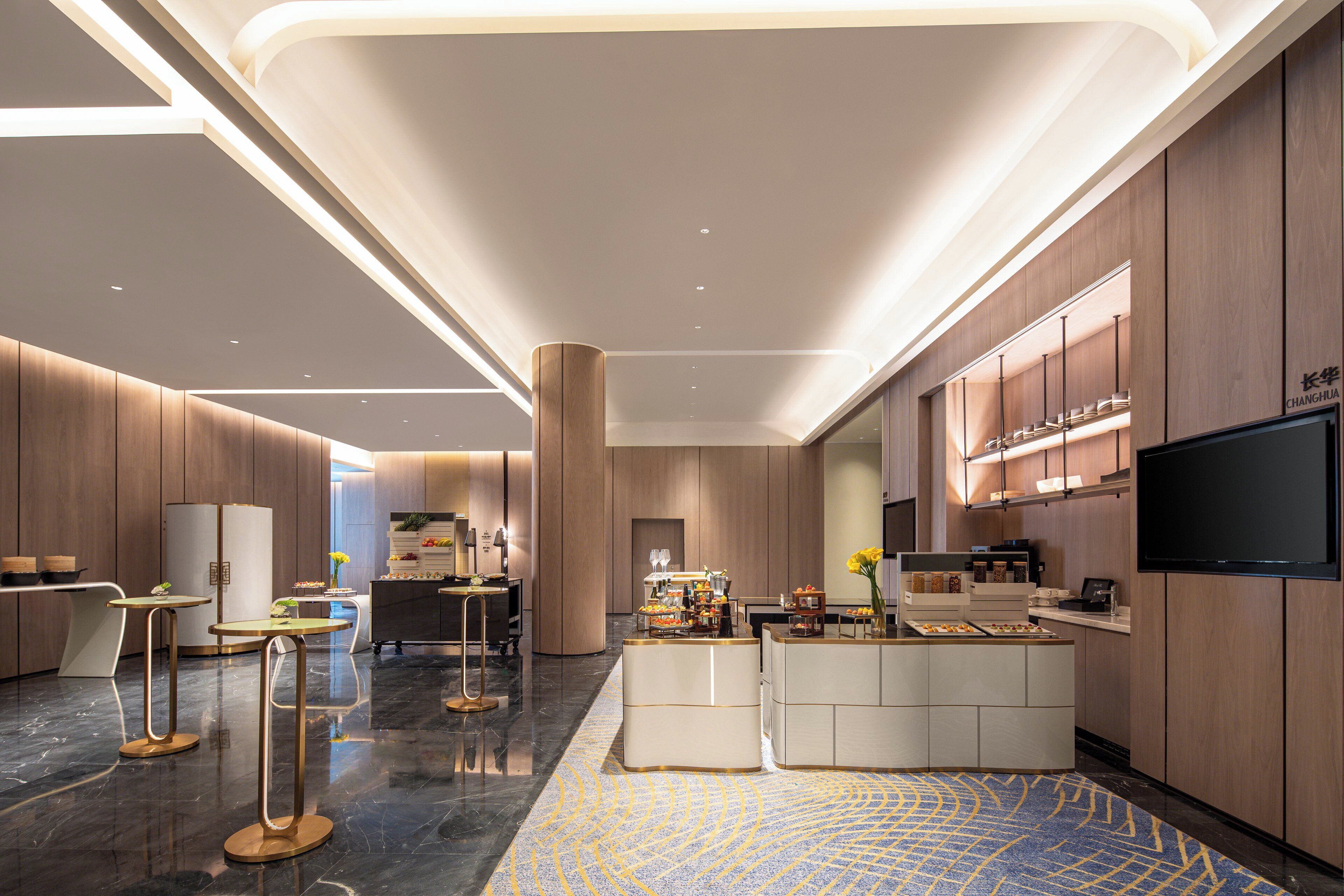 A modern hotel lobby with a central reception desk, a flat-screen TV, and a seating area with tables and chairs.