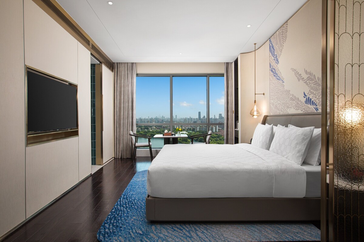 Habitación de hotel con una cama grande, televisión, balcón con vista a la ciudad y un detalle decorativo en la pared.