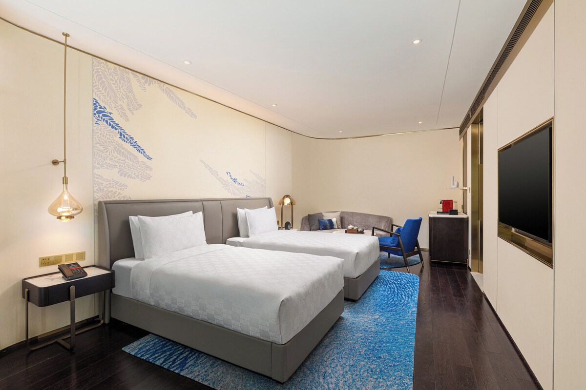 Habitación de hotel con dos camas, un escritorio, una silla, un televisor y un ventanal grande.
