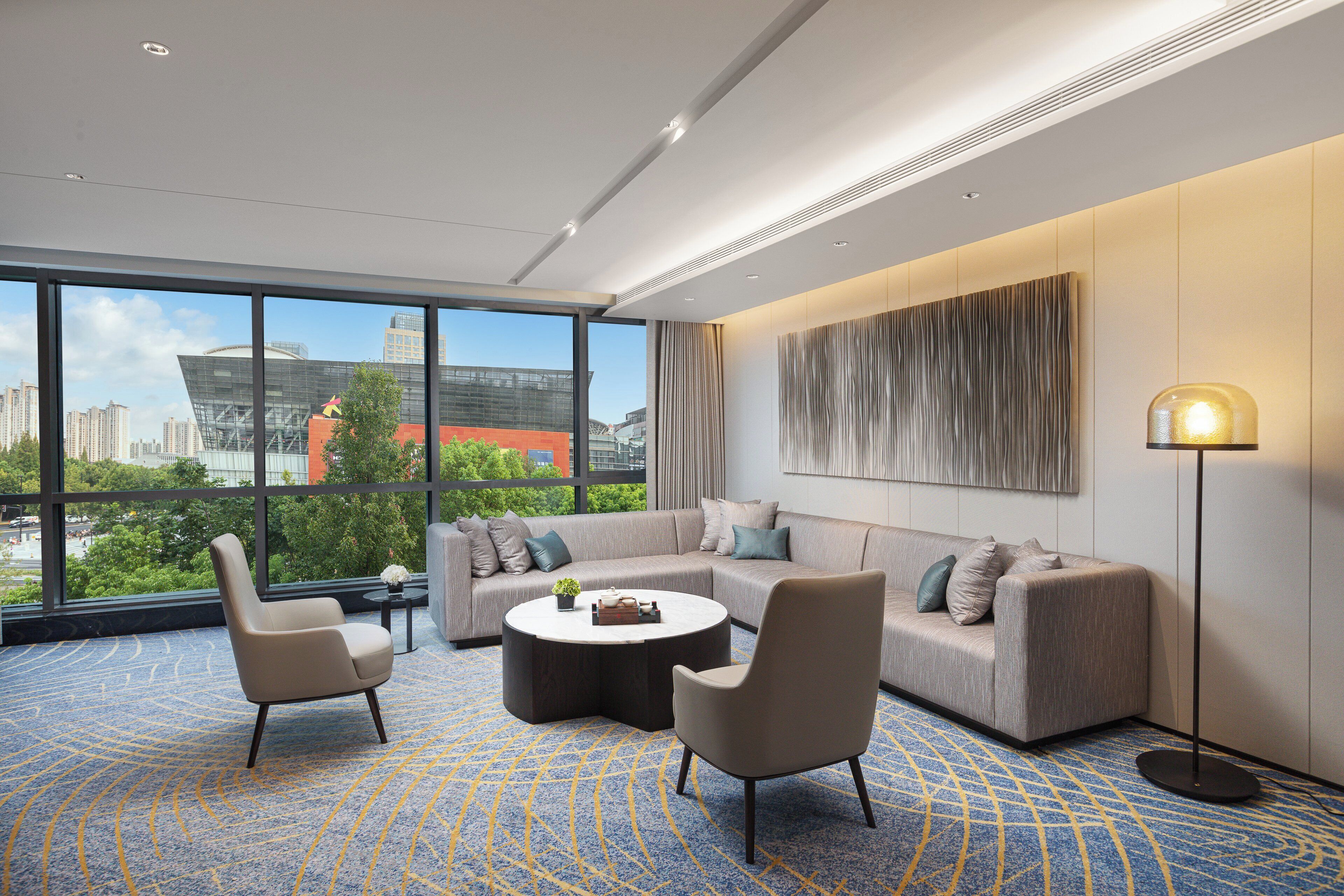 Una habitación de hotel moderna con un ventanal grande, un sofá, dos sillones, una mesita de centro y una lámpara de pie.