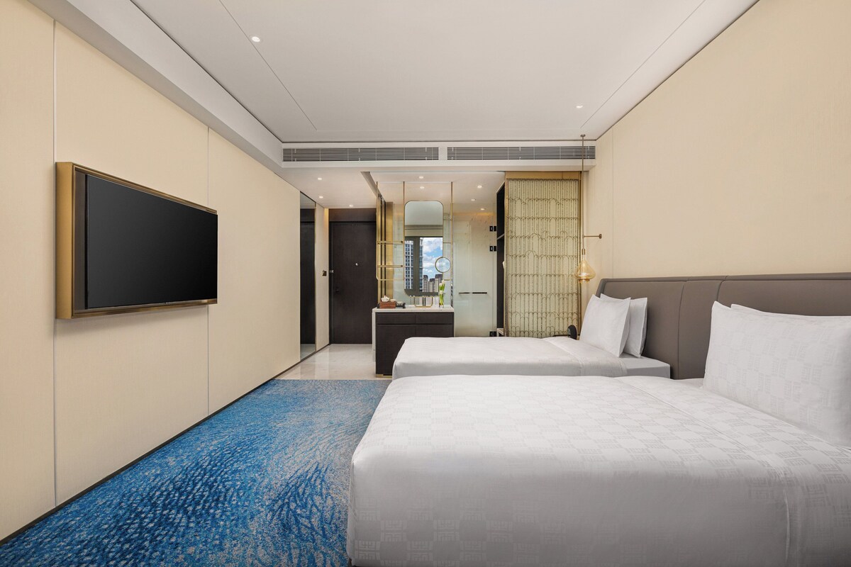 Habitación de hotel con dos camas, un televisor de pantalla plana, baño con espejo y vista a la ciudad.