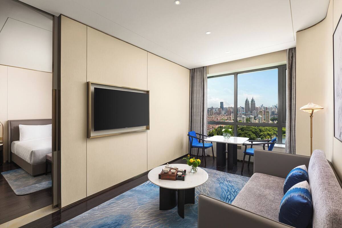 Ein modernes Hotelzimmer mit großem Fenster und Blick auf die Stadt, einem Flachbildfernseher an der Wand, einem Sofa, einem Esstisch mit Stühlen und einem kleinen Couchtisch.