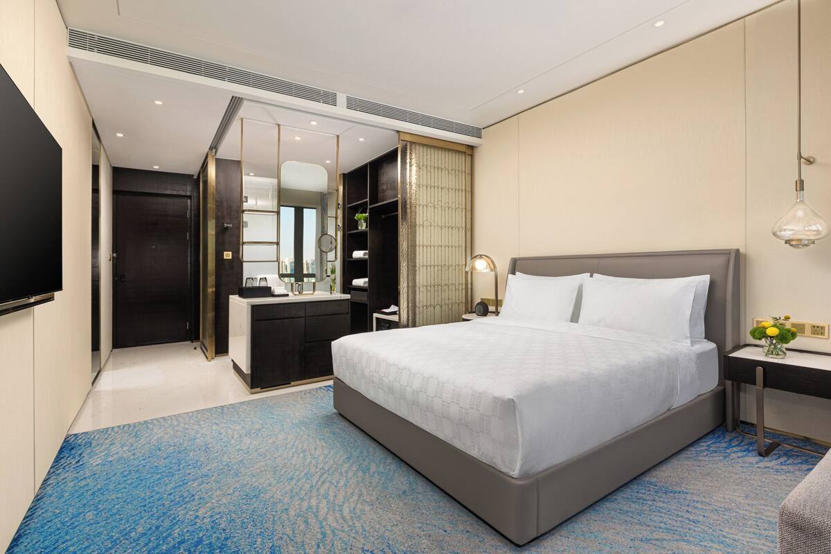 Ein modernes Hotelzimmer mit einem großen Bett, einem Nachttisch mit Lampe, einem Spiegel, einem Kleiderschrank und einem an der Wand befestigten Fernseher.