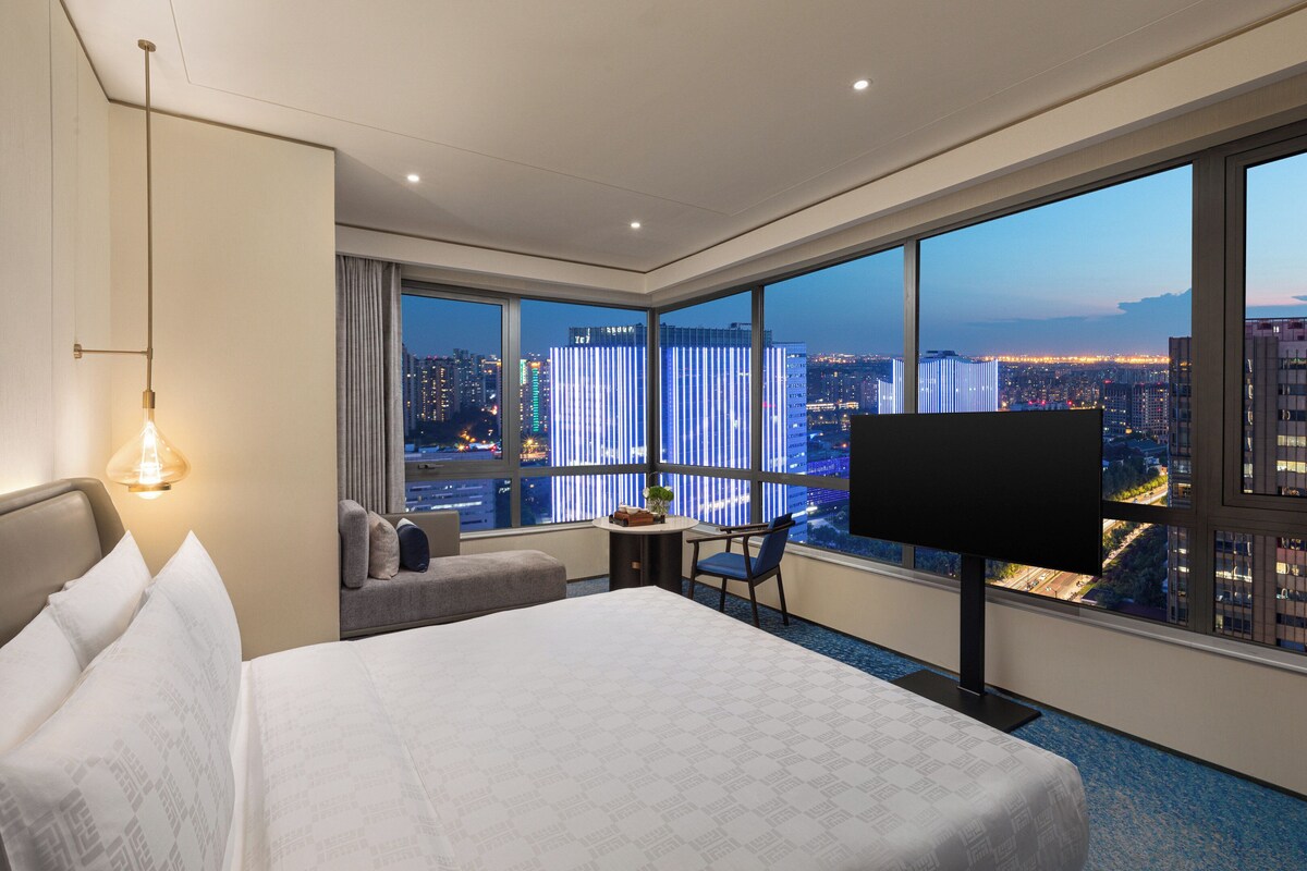 Habitación de hotel con una cama grande, un escritorio, una silla, un televisor y vistas a un paisaje urbano nocturno.