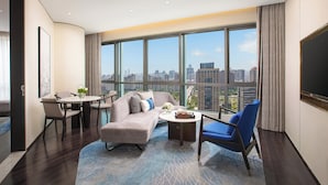 Una habitación de hotel moderna con un ventanal grande que ofrece vistas a la ciudad, un sofá, una mesa de comedor y un sillón azul.