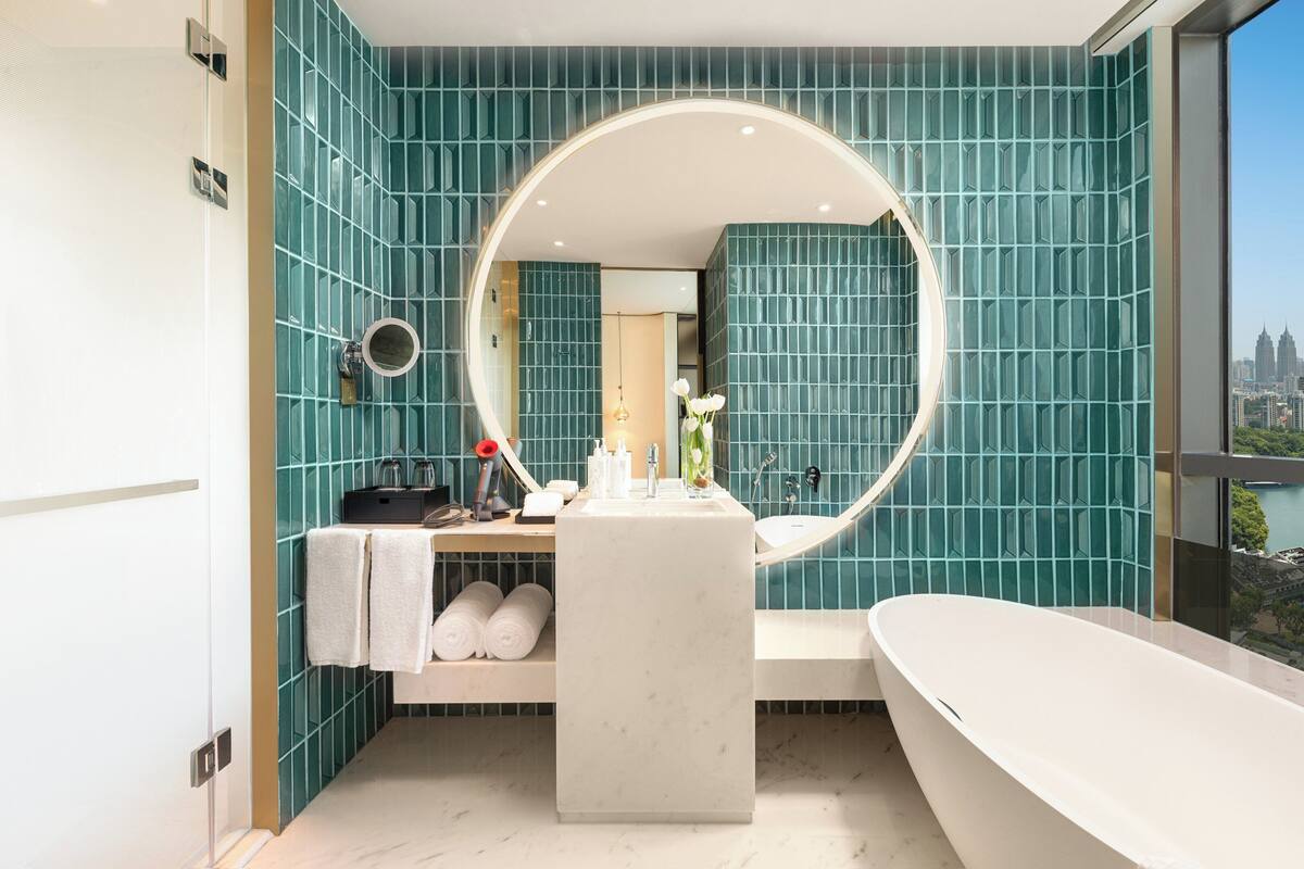 Ein modernes Badezimmer mit türkisfarbenen Fliesen, einer freistehenden Badewanne, einem großen Spiegel und Blick auf die Stadt.