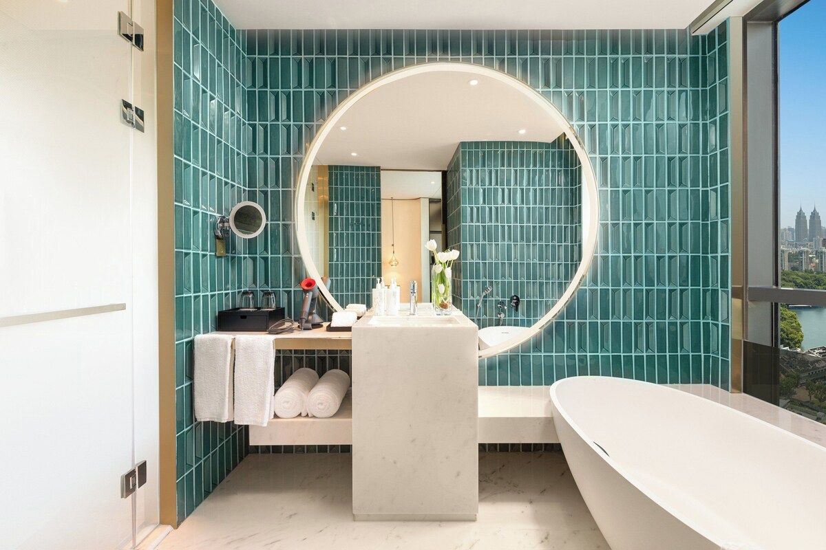 A modern bathroom with teal tiles, a freestanding bathtub, a large mirror, and a view of the city.