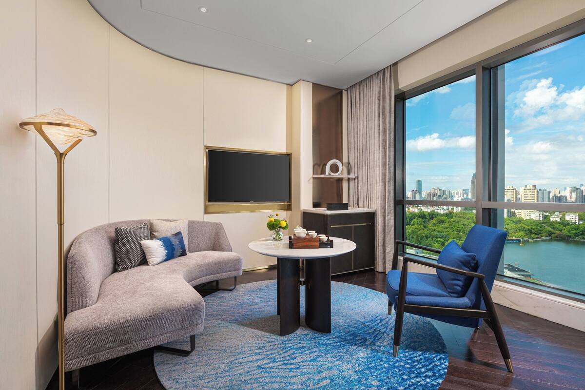 Ein modernes Hotelzimmer mit einer Couch, einem Sessel, einem runden Tisch, einem Fernseher und Blick auf die Stadt.