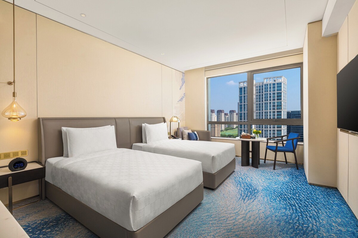 Habitación de hotel con dos camas, un escritorio con silla, televisión y un ventanal con vista a la ciudad.
