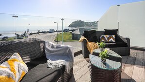 En solig uteservering med möbler i rotting och ett bord i glas, med utsikt över en kustväg och en strand.