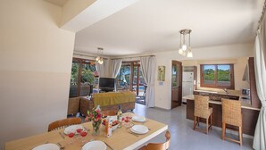 Un comedor amplio con una mesa dispuesta para cuatro, una cocina con ventana, y una sala de estar con televisor y asientos exteriores.