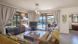 Amplia sala de estar con sofá, una mesa de centro y televisor. Puerta corrediza de vidrio que da a un área exterior con piscina.