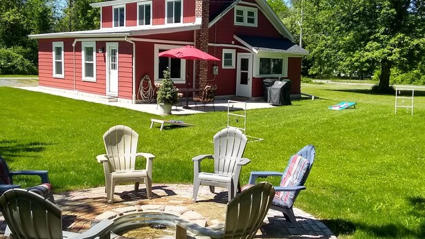 A two-story house with a red exterior and white trim, a brick chimney, and a patio with Adirondack chairs and a red umbrella.