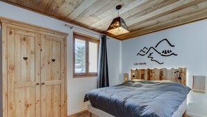 Slaapkamer met een houten garderobe, een bed met een grijze dekbedovertrek, een raam met uitzicht op bomen, en een muur met een bergtekening en de tekst 'Les Nards'.