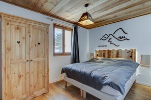 A bedroom with a wooden wardrobe, a bed with a grey duvet, a window with a view of trees, and a wall with a mountain drawing and the text "Les Nards".