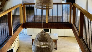 A spacious loft with exposed wooden beams, a balcony with a railing, and a hanging woven light fixture.