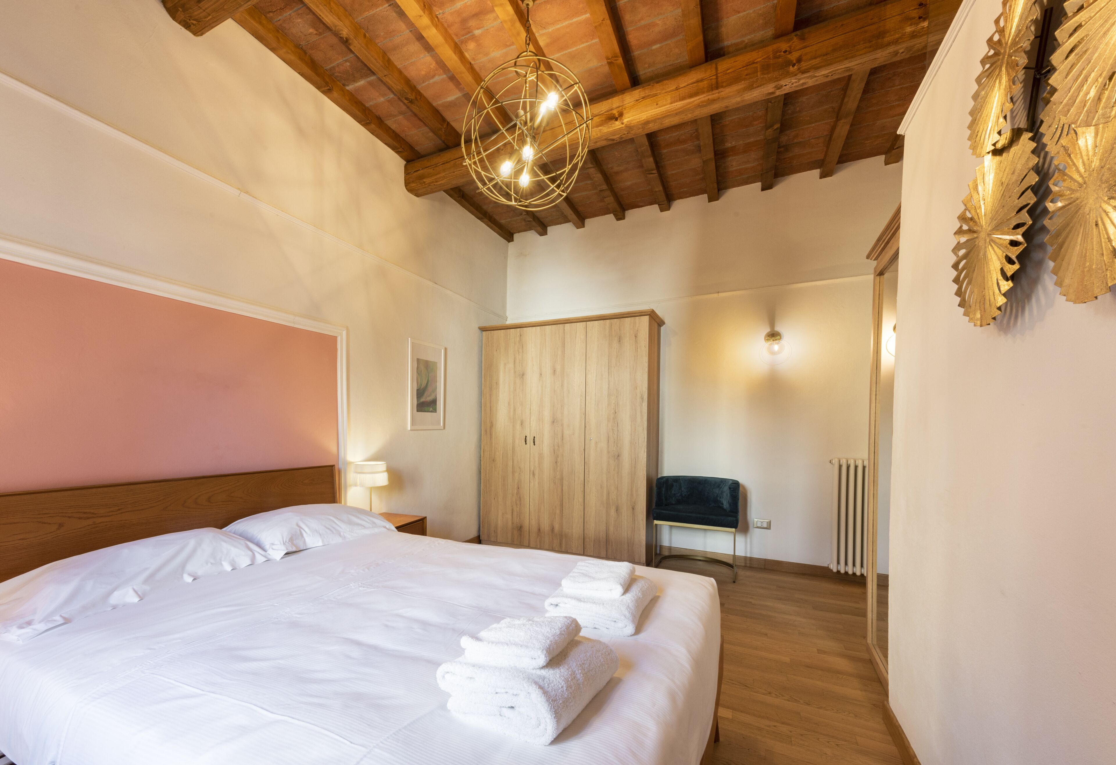 A hotel room with a large bed, wooden ceiling beams, a wooden wardrobe, a chair, and a radiator.