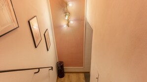 Una escalera estrecha con piso de madera, un bote de basura negro y una lámpara fijada a la pared.