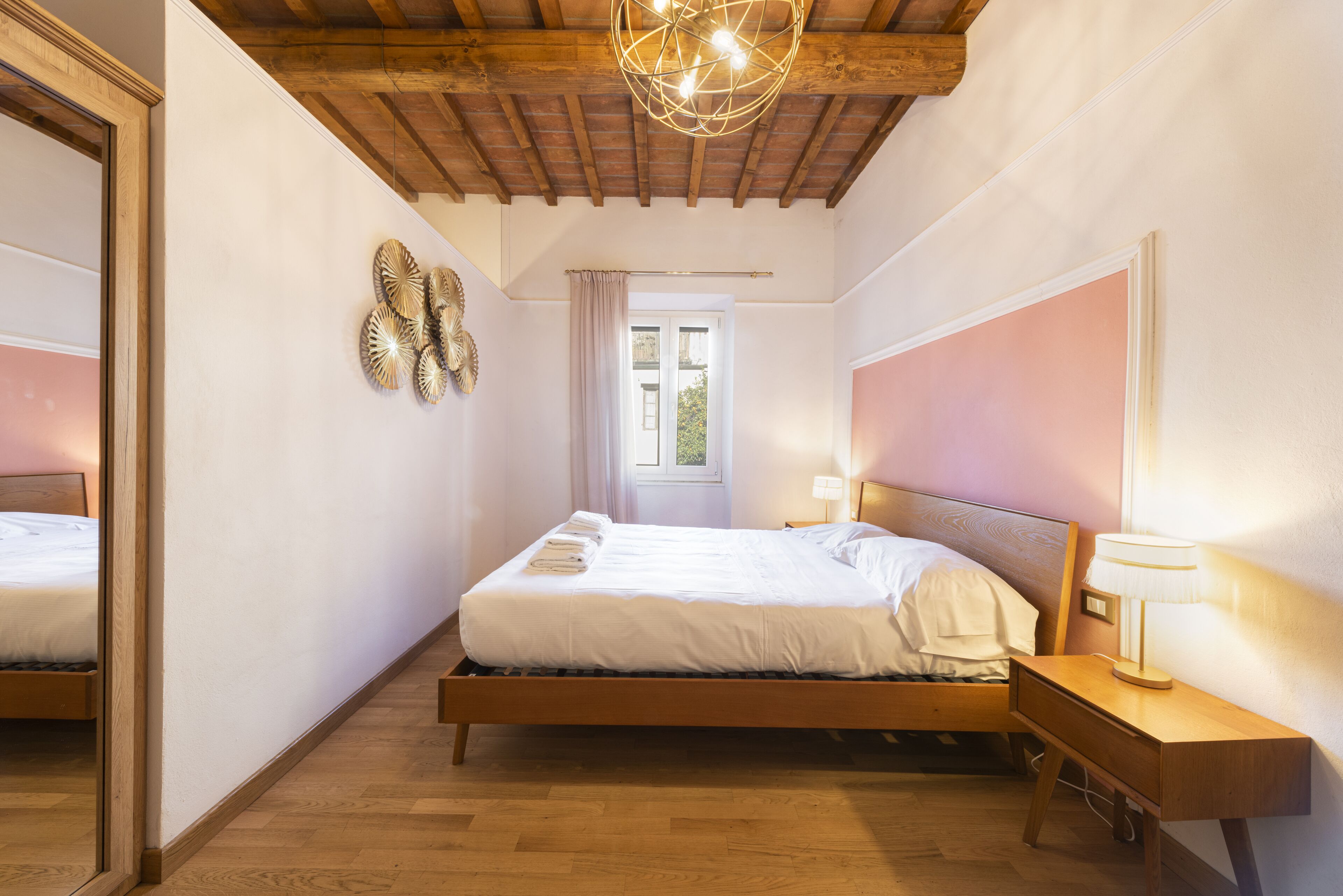 A bedroom with a wooden bed, a nightstand with a lamp, a mirror, and a window with a view of greenery.