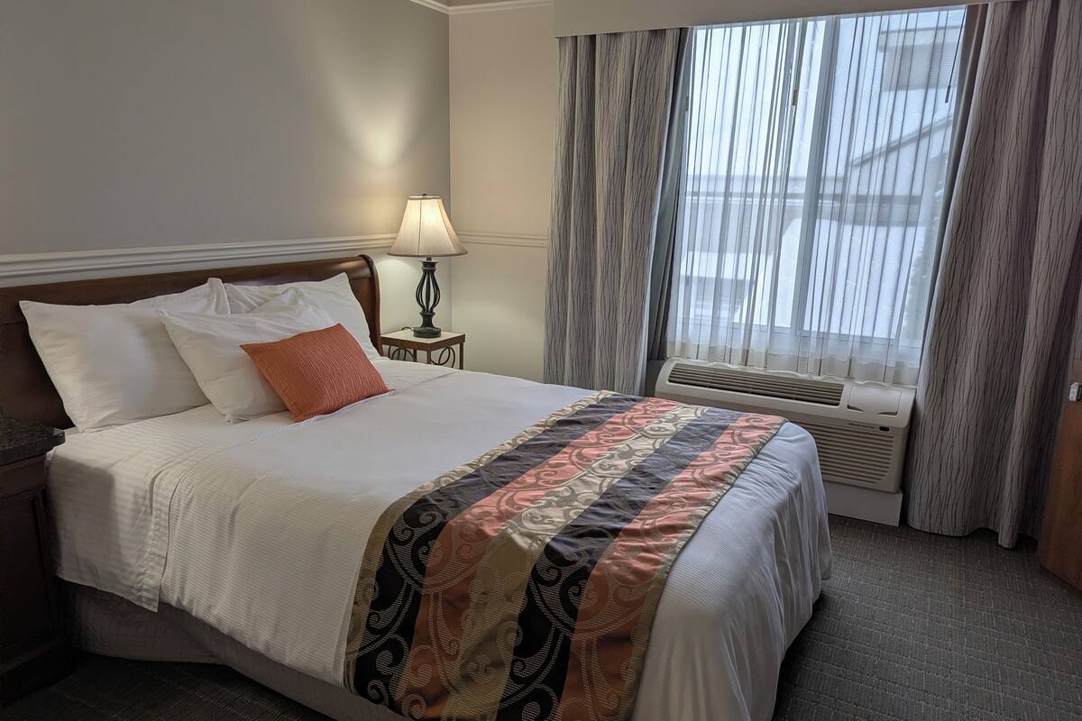A hotel room with a bed, a nightstand with a lamp, a window with curtains, and an air conditioning unit.