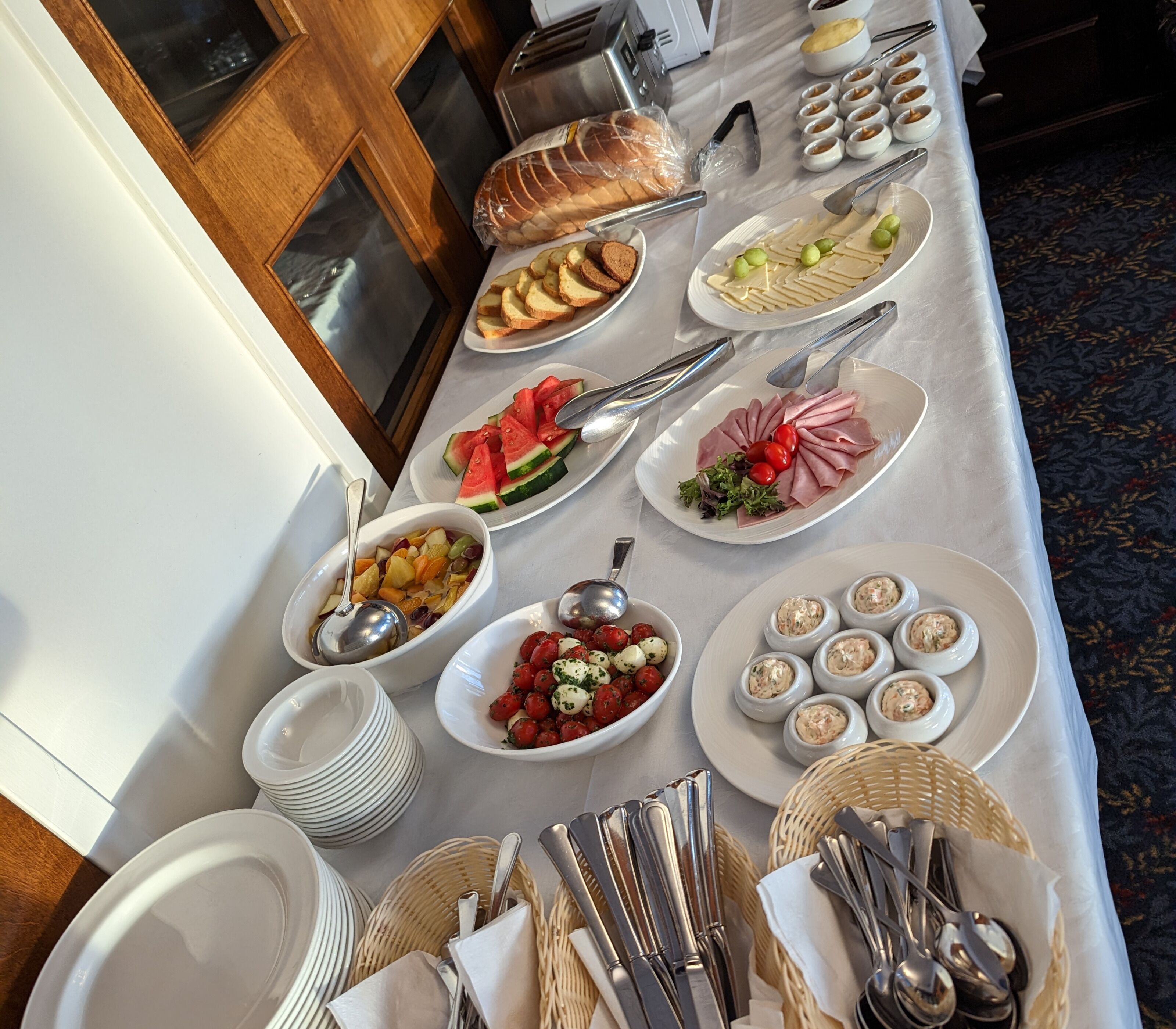 Una mesa de buffet con diversos alimentos, entre ellos frutas, quesos y carnes, además de utensilios y platos para servir.
