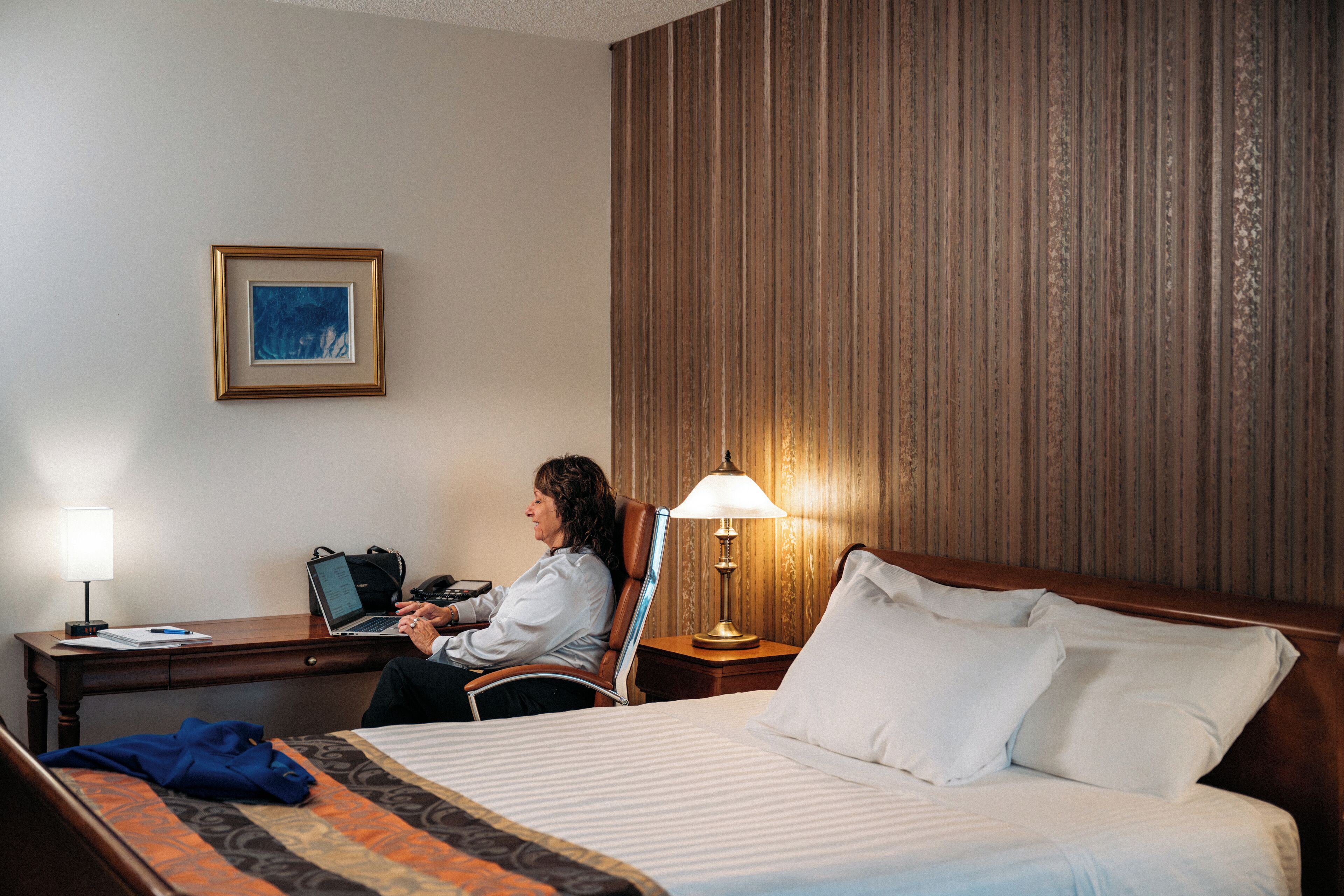 Habitación de hotel con cama, escritorio con una computadora portátil, lámpara y un cuadro en la pared.