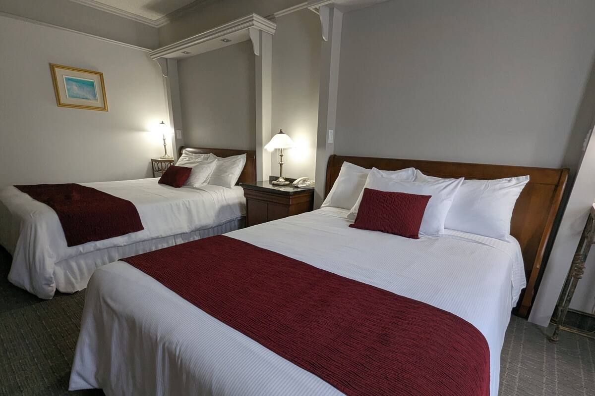 Two double beds with white linens and red throw blankets, a nightstand with a lamp, and a framed picture on the wall.