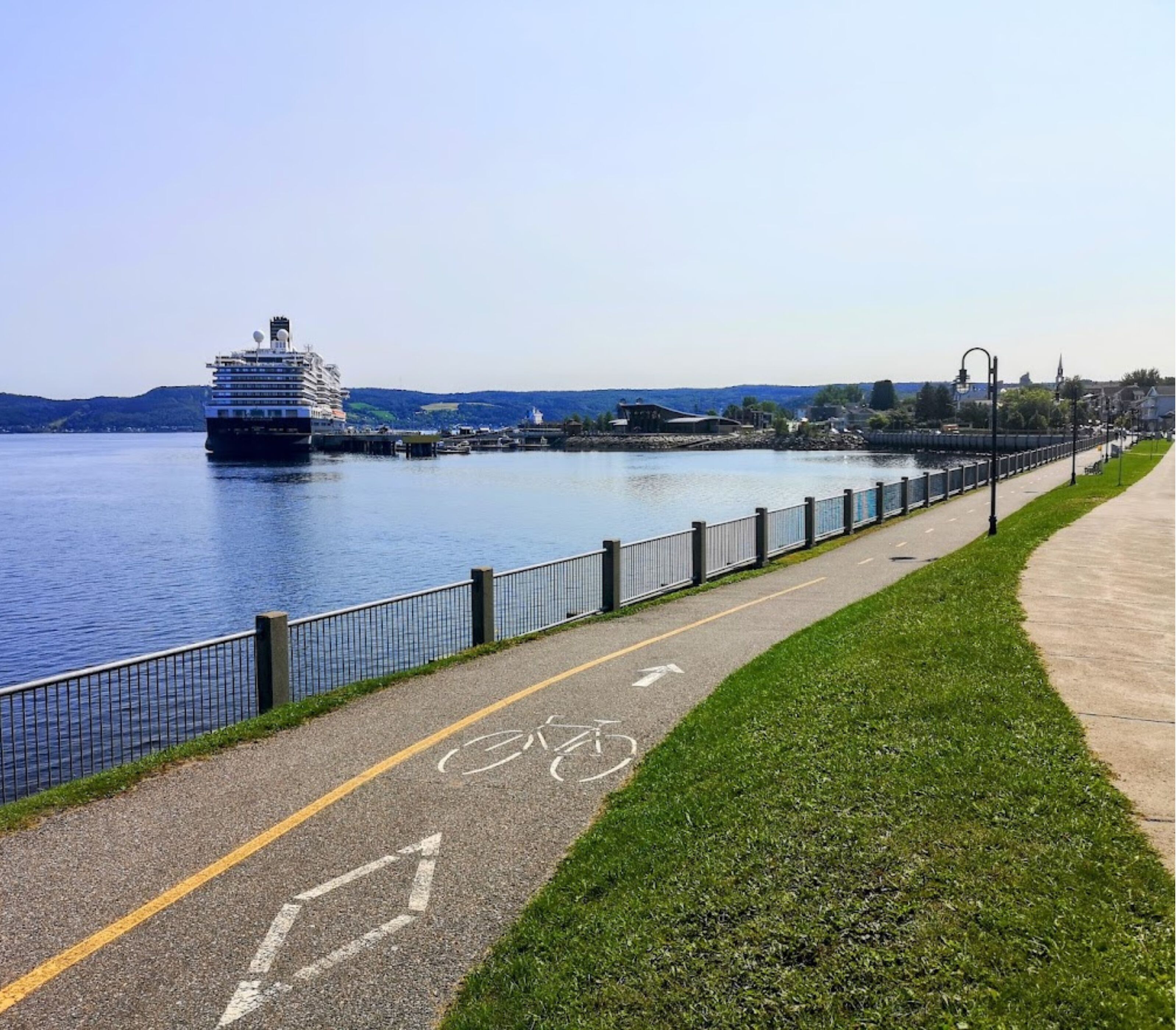 A waterfront promenade with a bike lane, a cruise ship docked, and a cityscape in the background.