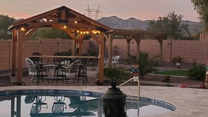 Un patio trasero con una pérgola de madera, mobiliario exterior, una piscina y estatuas decorativas.