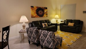 Un salón con un sofá de cuero negro, un reposapiés estampado y una alfombra amarilla. Hay dos lámparas sobre mesitas auxiliares, una obra de arte colgada en la pared con un girasol y una puerta con un perchero.