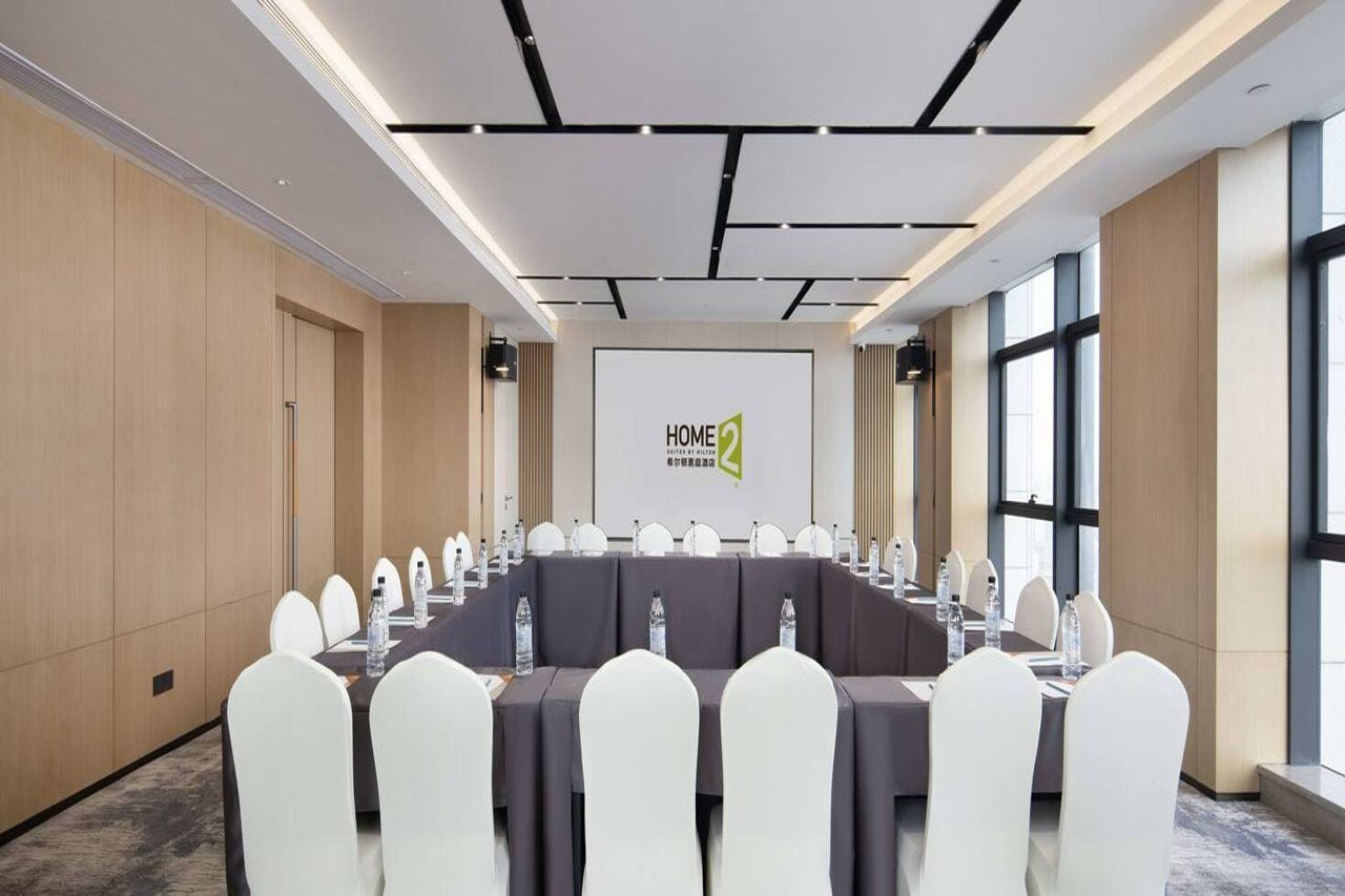A conference room with a long table, white chairs, and a screen displaying 'HOME2' branding.
