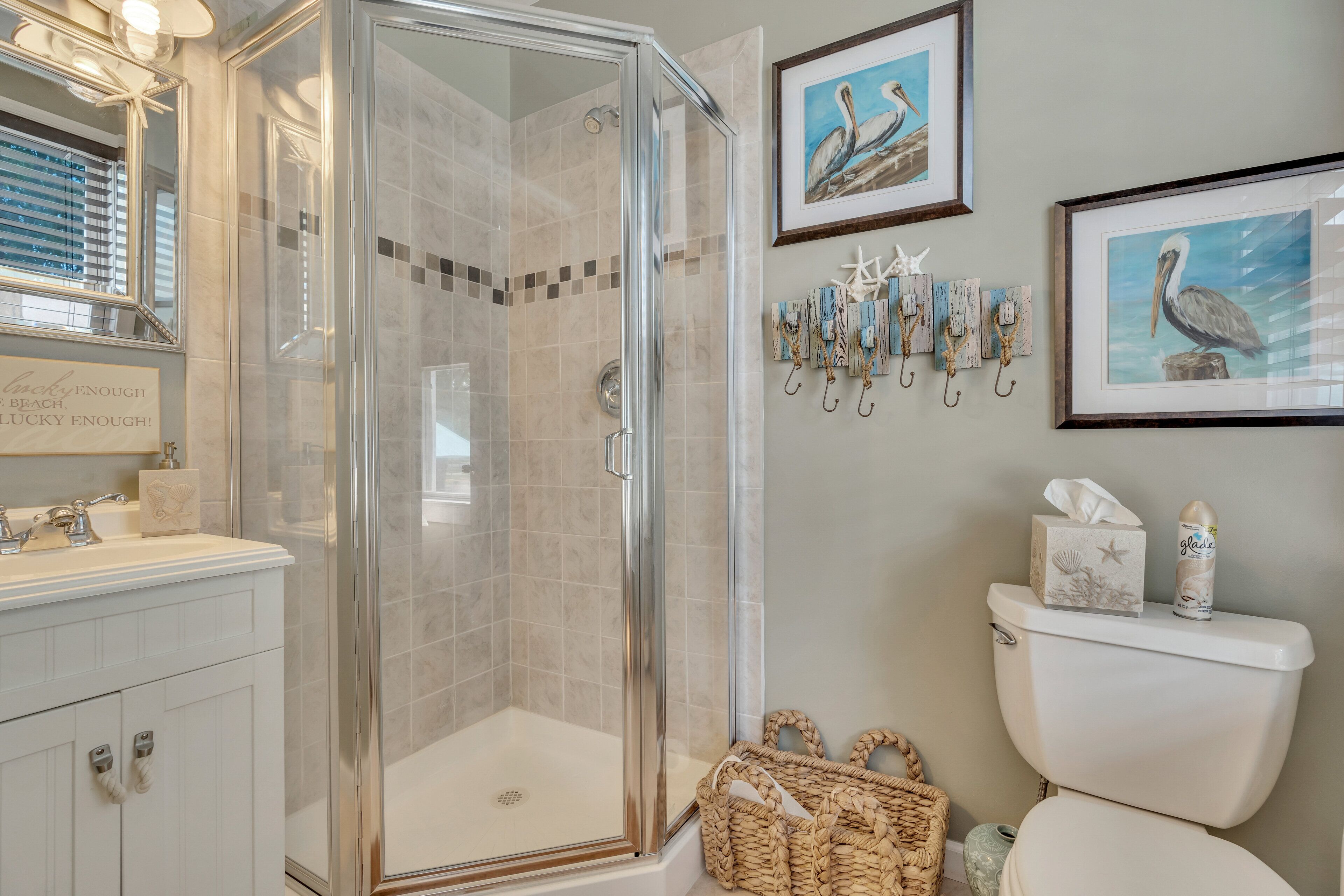 A bathroom with a glass-enclosed shower, a white sink, and a toilet. There are framed pictures on the wall and a basket on the floor.