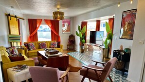 Un salon avec un canapé jaune, une table basse et un téléviseur. On y trouve deux fauteuils, un tapis et une plante.