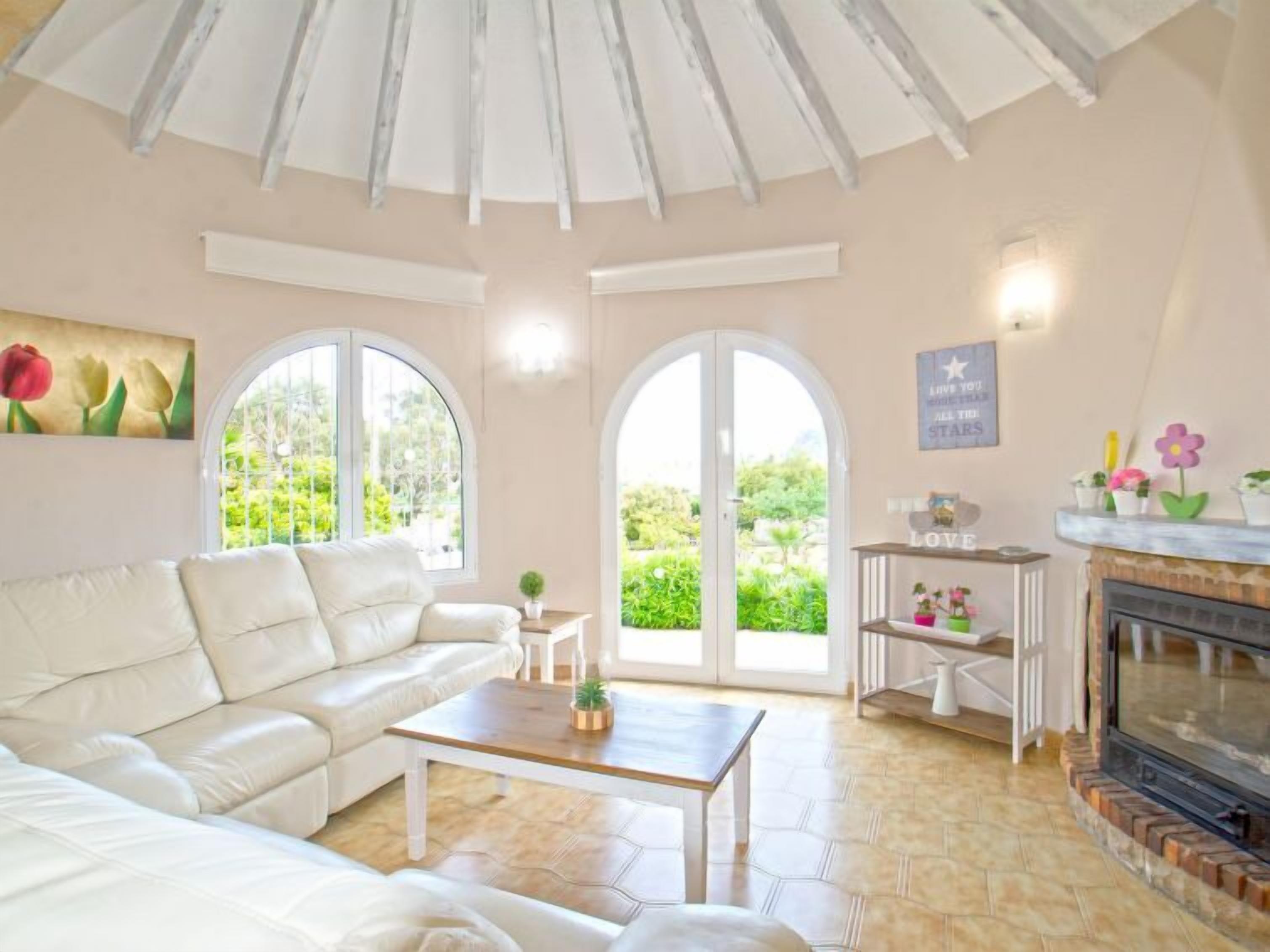 Ein großzügiges Wohnzimmer mit einem weißen Ledersofa, einem Holztisch und einem Kamin. Große Fenster bieten Blick ins Grüne.