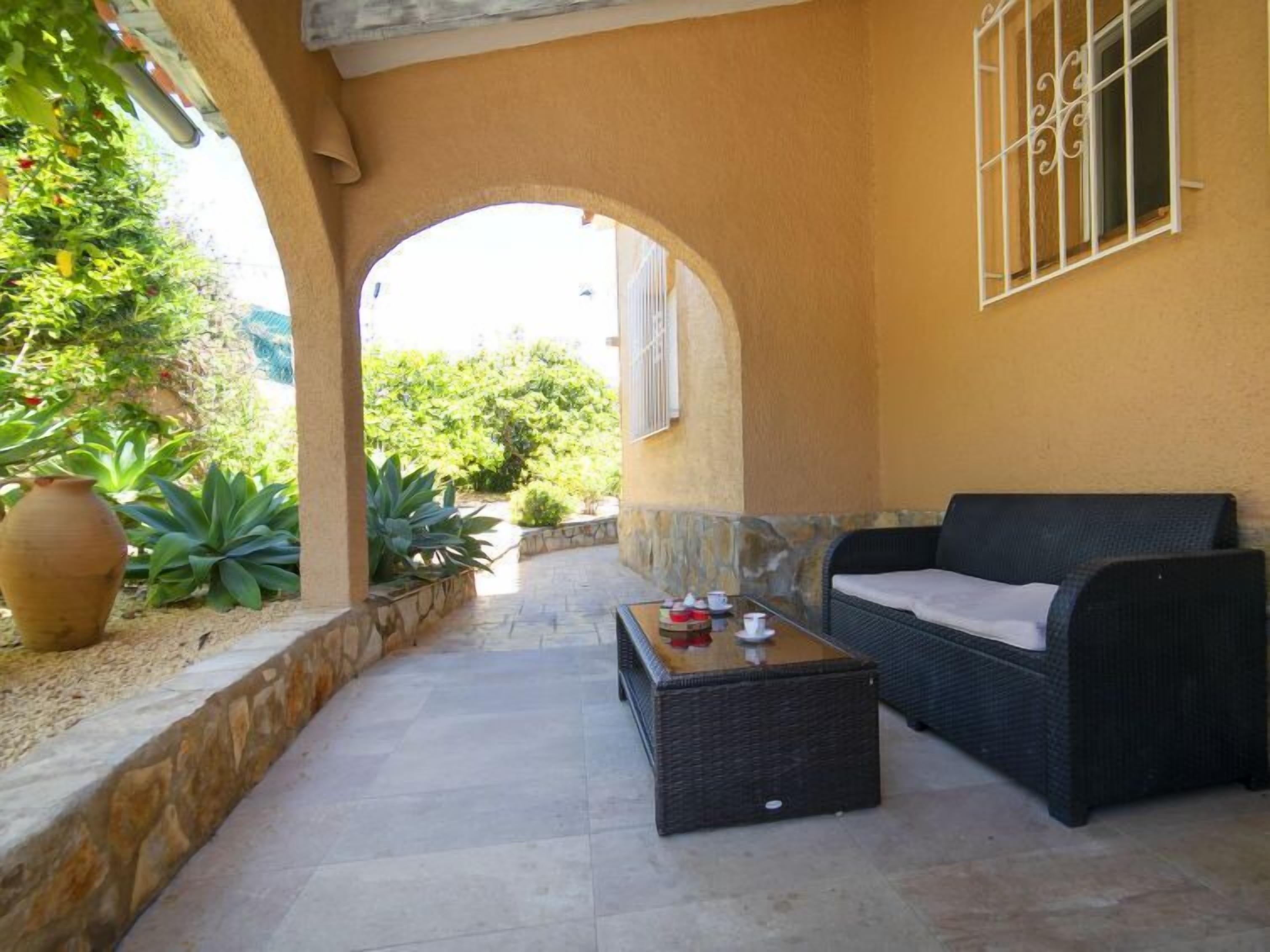 Patio esterno con divano in vimini e un tavolino, circondato da piante e un muro in pietra.