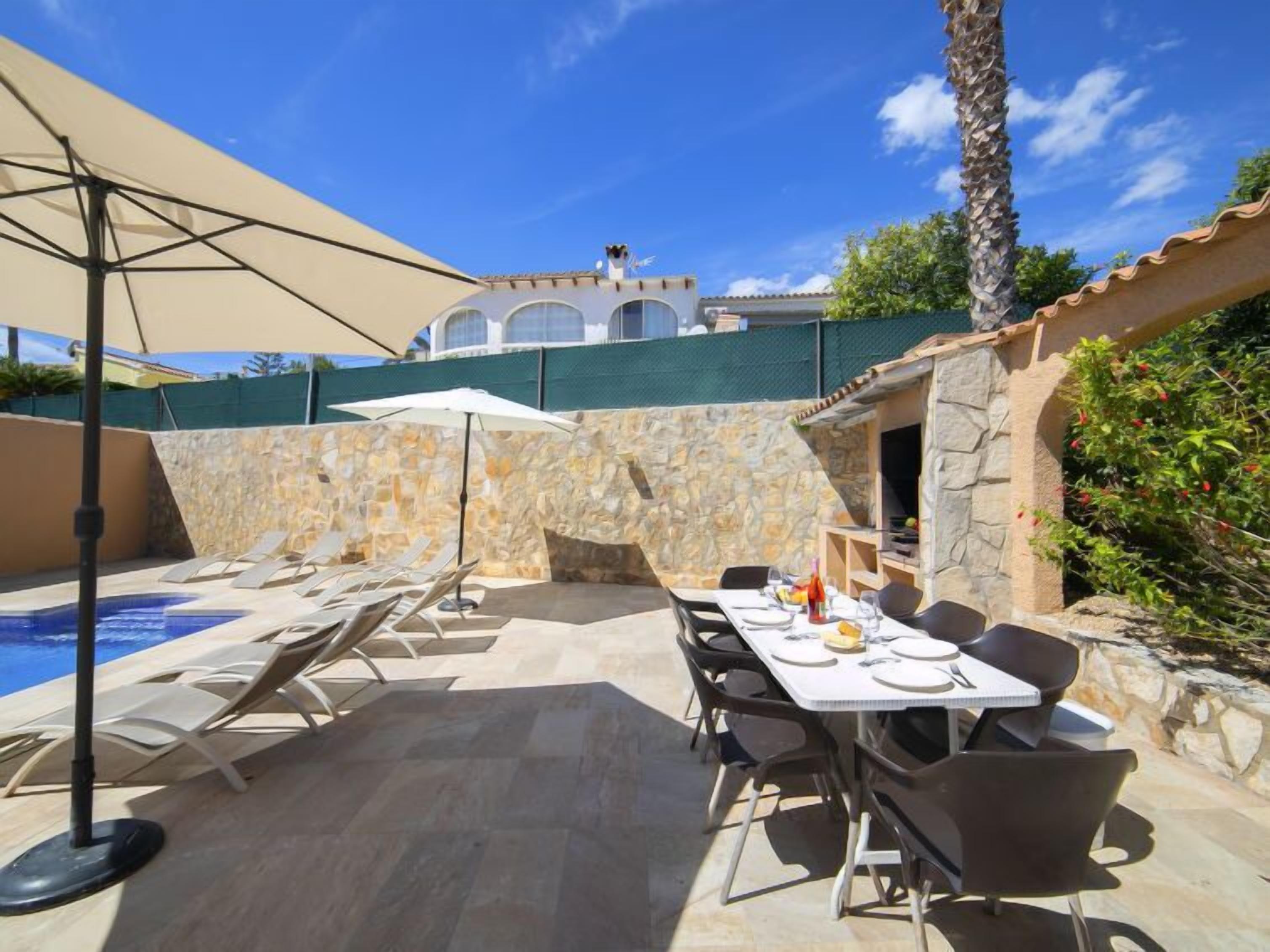 Zona de piscina con sillones de descanso, una mesa preparada para comer y un muro de piedra de fondo.