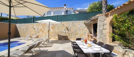 Zona de piscina con sillones de descanso, una mesa preparada para comer y un muro de piedra de fondo.