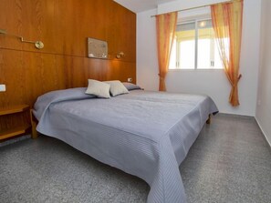 A bedroom with a wooden headboard, a bed with a grey bedspread, two pillows, a window with orange curtains, and a small bedside table.